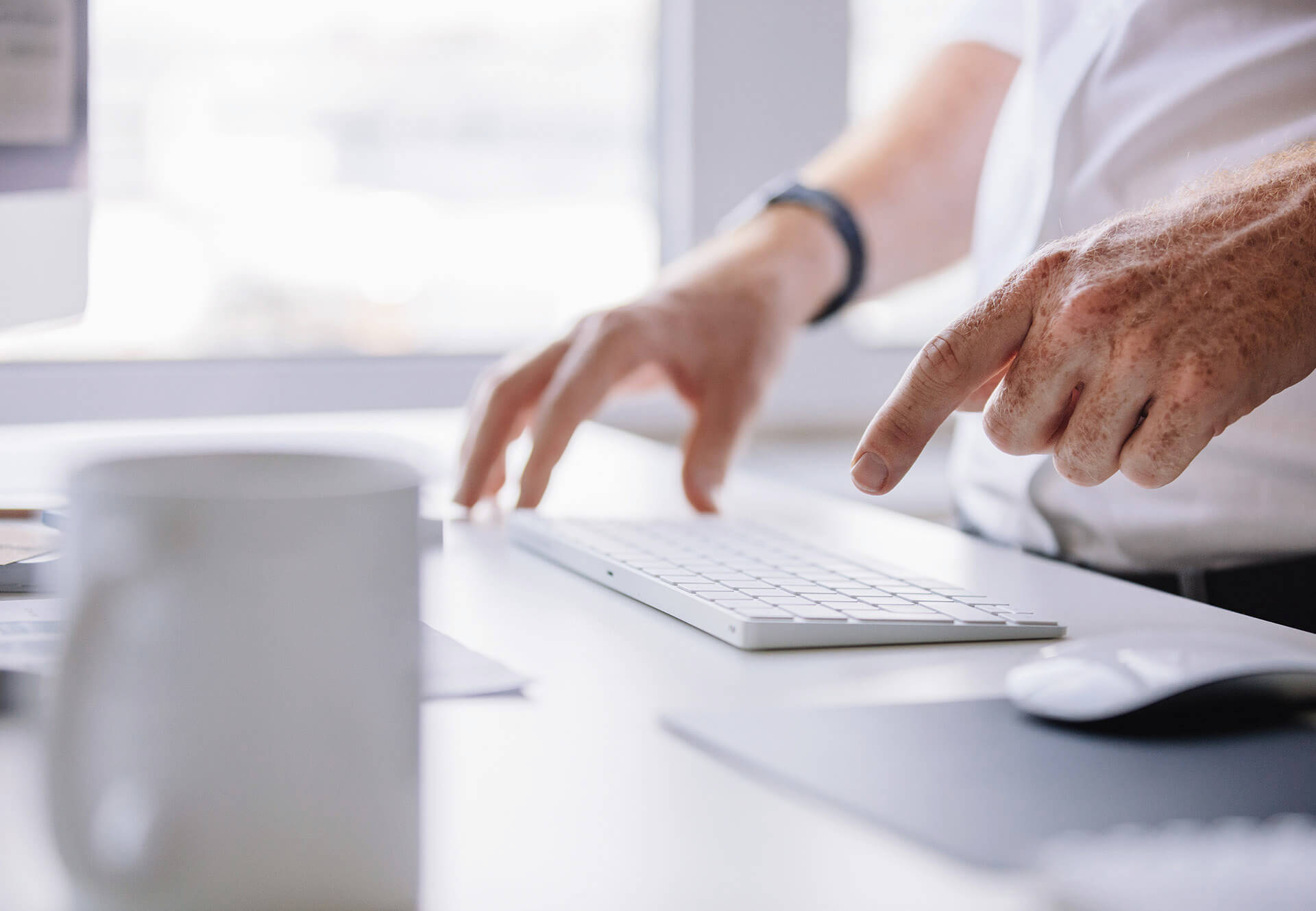 Two persons hands are hovering above a keyboard a coffee mug is shown blurred in the front of the pciture